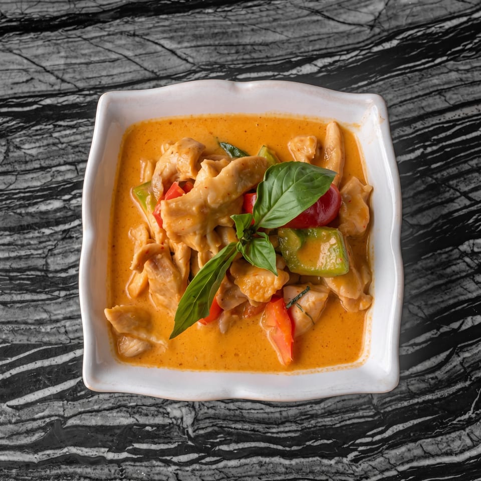 Red curry with chicken, bell peppers, and basil in a white bowl on a marbled table.