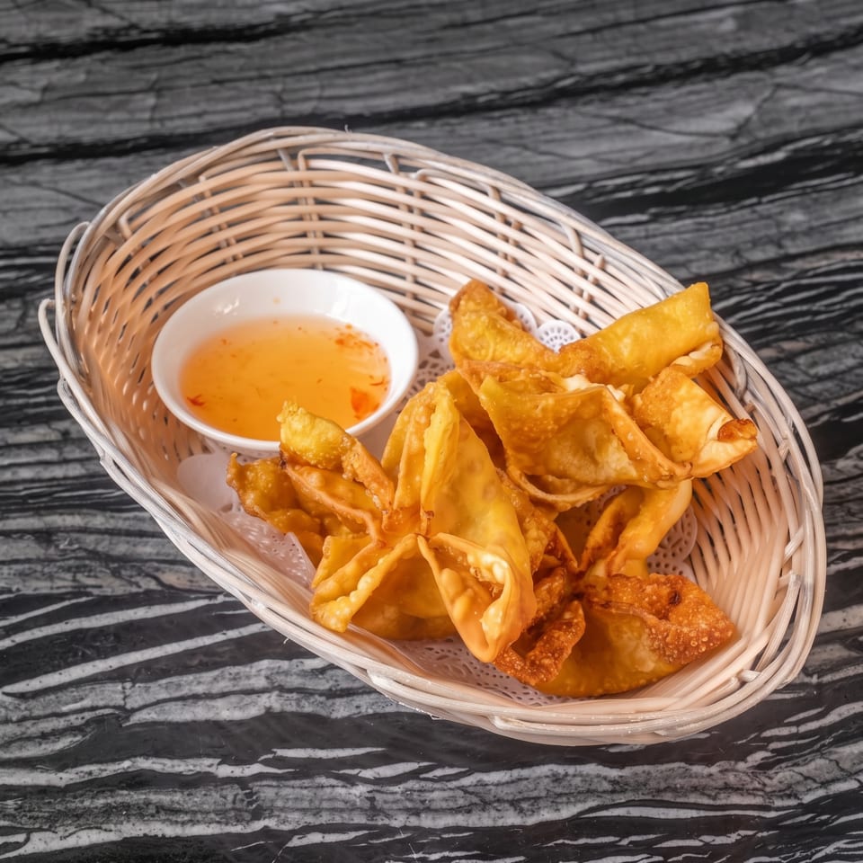 Crispy golden crab rangoons in a basket with sweet chili dipping sauce on a dark marble surface.