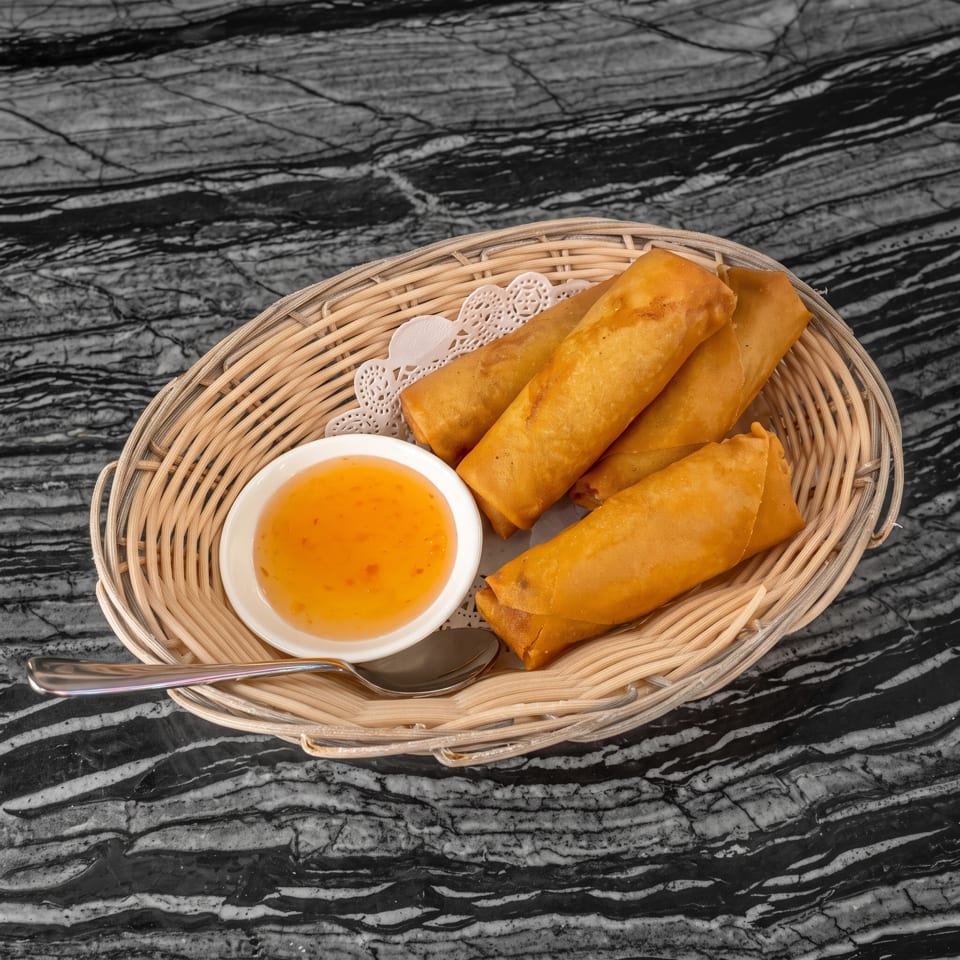 Golden-brown fried spring rolls in a wicker basket with a side of orange dipping sauce and a spoon.