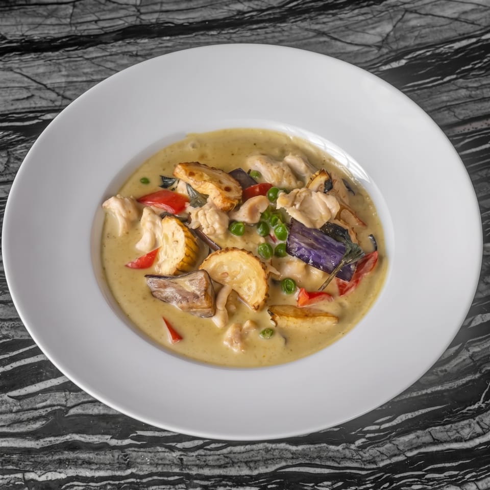 Yellow-green curry with chicken, eggplant, red peppers, and peas in a white bowl on a marble table.